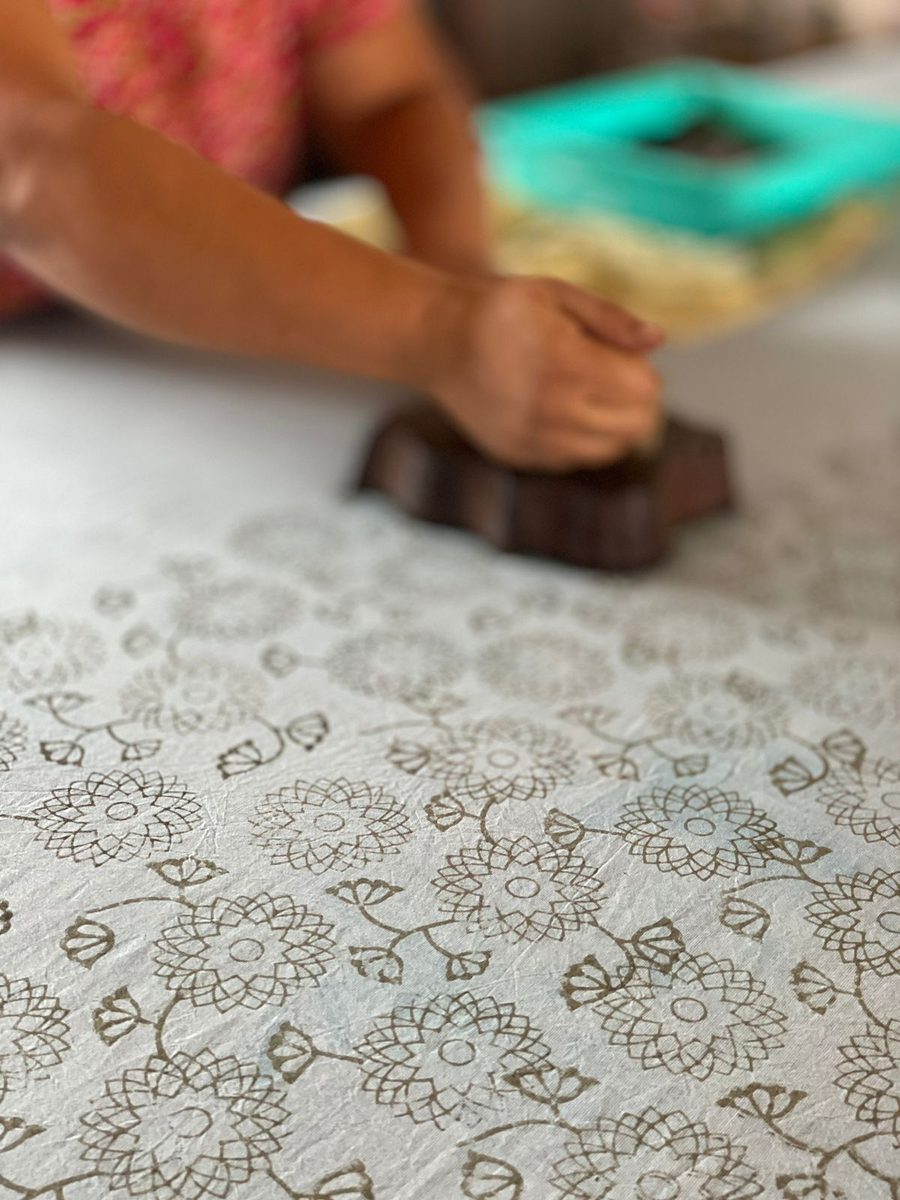 A hand pressing a carved wooden block onto white cotton, leaving a grey floral print